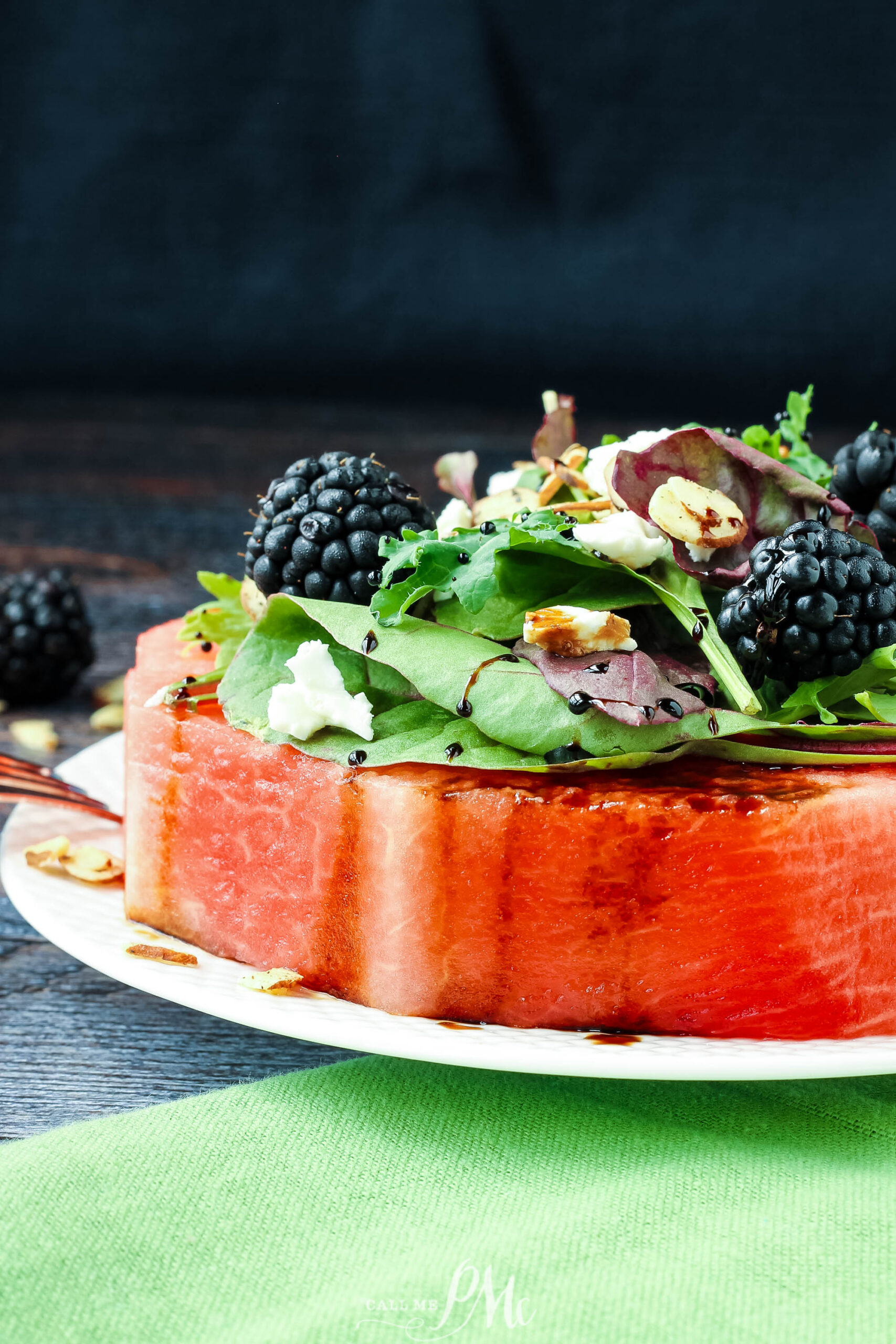 A thick slice of watermelon topped with mixed greens, blackberries, feta crumbles, almond slices, and balsamic glaze—this Feta Almond Watermelon Salad is served fresh on a white plate.