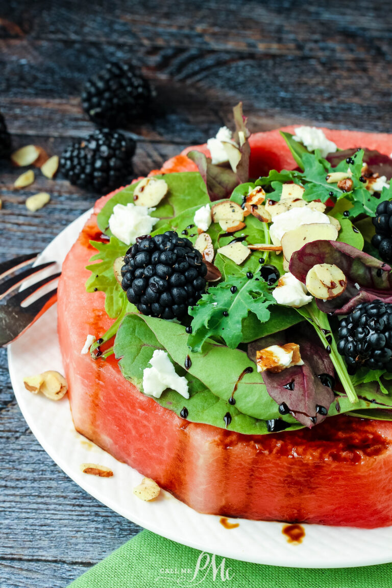 A thick slice of watermelon serves as the base for a vibrant Feta Almond Watermelon Salad, topped with mixed greens, blackberries, sliced almonds, feta cheese, and balsamic glaze, presented on a white plate with a fork beside it.