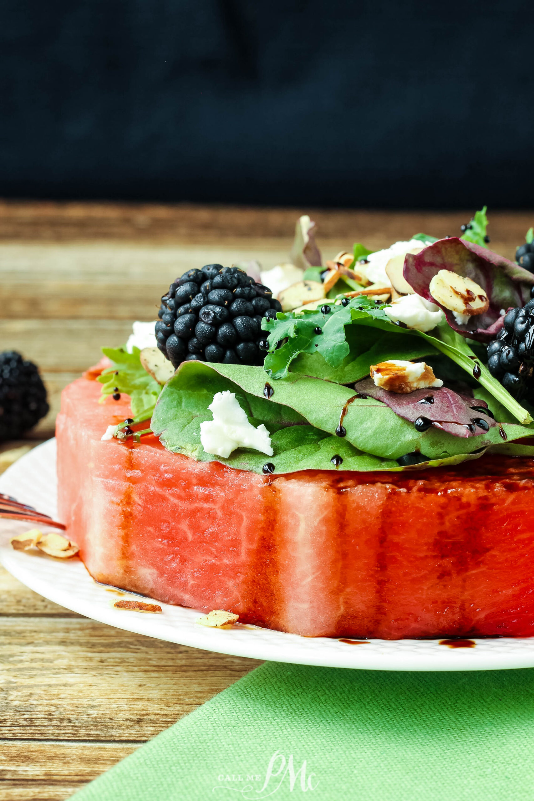 A thick slice of watermelon becomes the base for this Feta Almond Watermelon Salad, topped with mixed greens, blackberries, crumbled feta, sliced almonds, and a drizzle of balsamic glaze on a white plate against a wooden background.