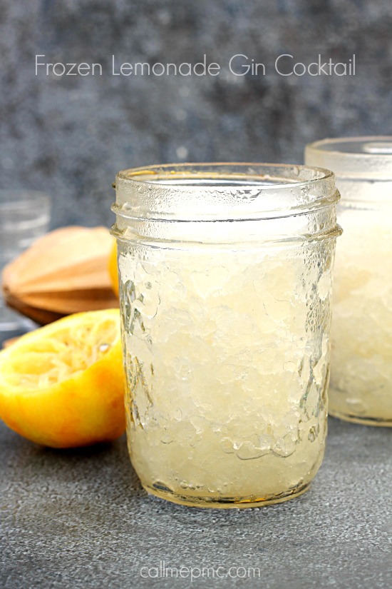 A mason jar filled with a Frozen Lemonade Gin Cocktail sits on a gray surface, with a halved lemon and a citrus juicer in the background.
