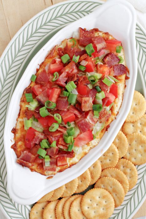 Oval dish topped with chopped bacon, tomatoes, and green onions, served with round crackers on a large plate.