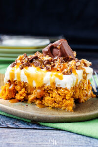 A slice of Skinny Pumpkin Snickers Poke Cake with a crumbly orange base, topped with whipped cream, caramel drizzle, chopped chocolate, and a chocolate bar piece, served on a green plate with a fork nearby.