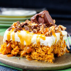 A slice of Skinny Pumpkin Snickers Poke Cake with a crumbly orange base, topped with whipped cream, caramel drizzle, chopped chocolate, and a chocolate bar piece, served on a green plate with a fork nearby.