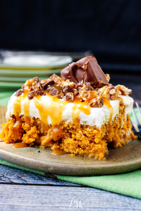 A slice of Skinny Pumpkin Snickers Poke Cake with a crumbly orange base, topped with whipped cream, caramel drizzle, chopped chocolate, and a chocolate bar piece, served on a green plate with a fork nearby.