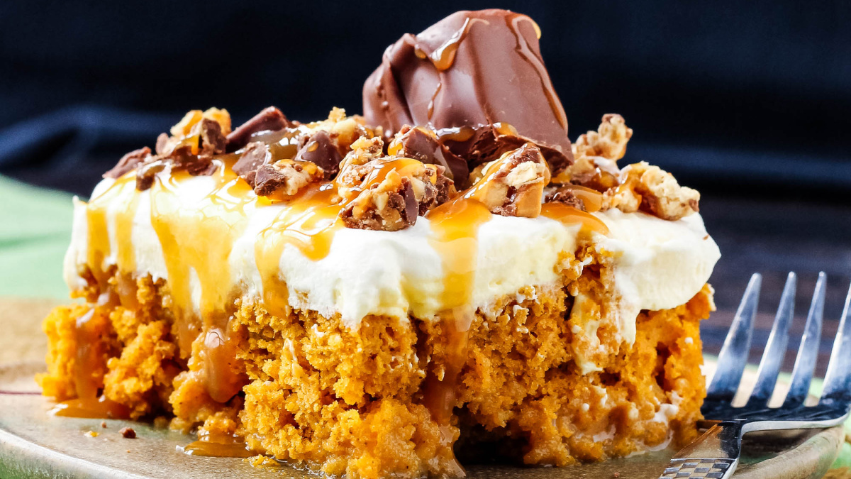 A close-up of a slice of Skinny Pumpkin Snickers Poke Cake topped with creamy frosting, caramel drizzle, chopped nuts, and a chocolate piece, placed on a plate with a fork nearby.