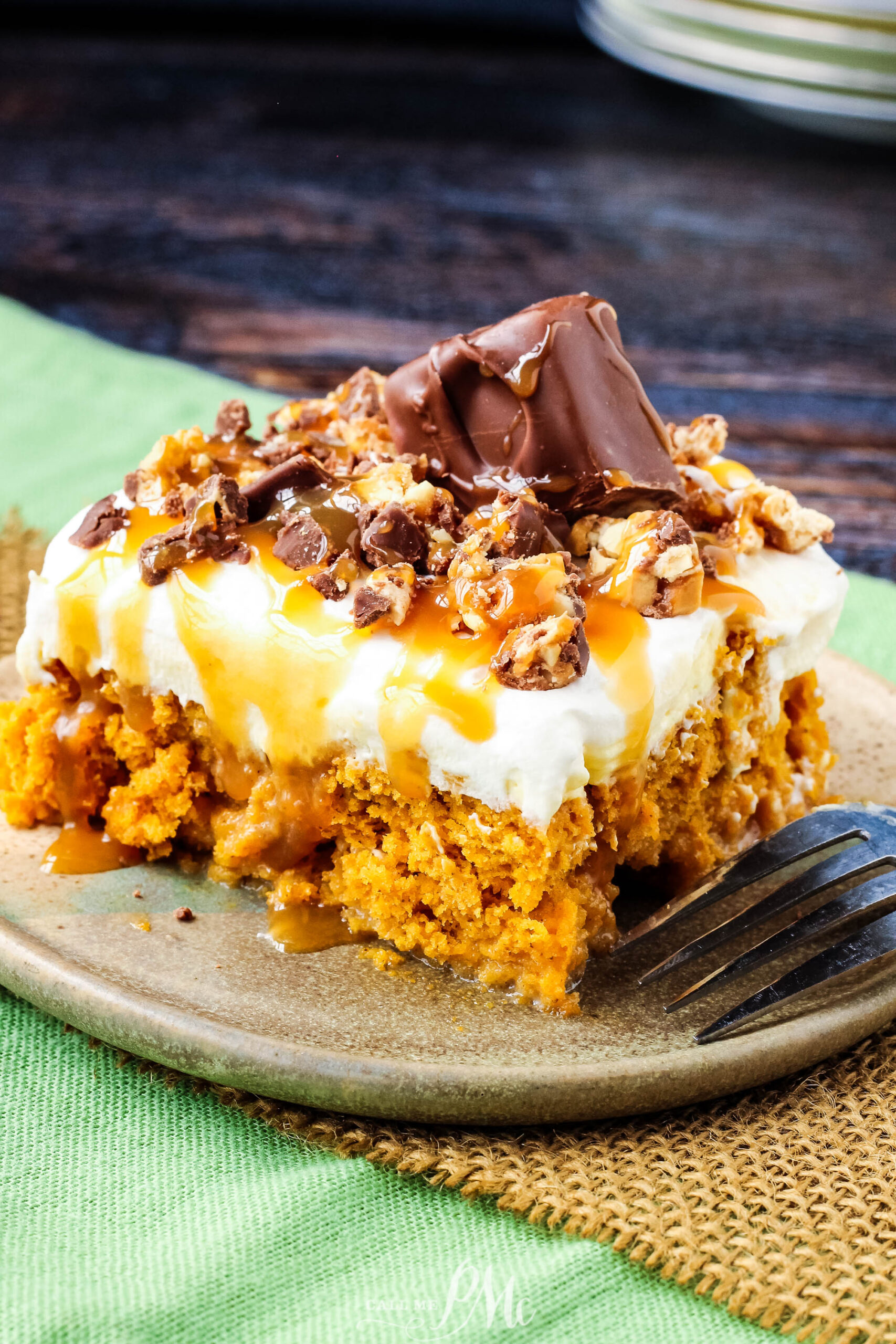 A slice of caramel cake topped with whipped cream, chopped chocolate pieces, and caramel drizzle sits on a plate with a fork beside it, reminiscent of the rich flavors found in a Skinny Pumpkin Snickers Poke Cake.