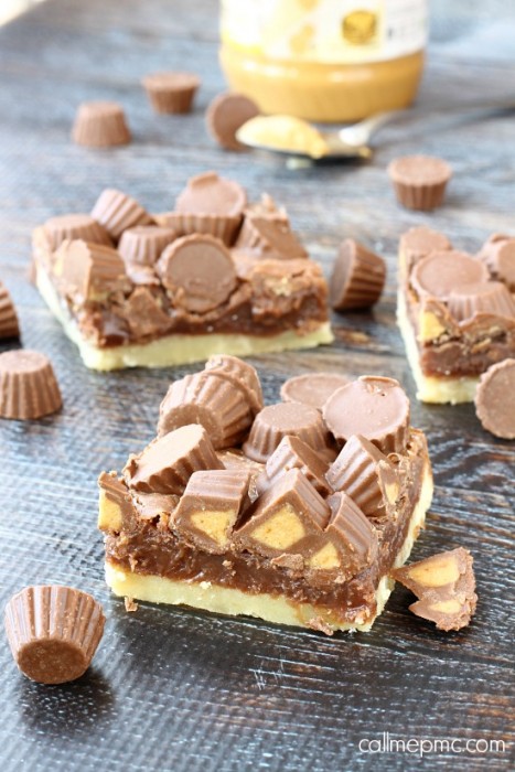 Buttery and flaky shortbread is covered with a rich chocolate peanut butter mixture then topped with peanut butter cup minis in these Chocolate Peanut Butter Shortbread Bars. Rich like fudge, this dessert is not for the faint of heart. Dessert topped with mini peanut butter cups are displayed on a wooden surface, with extra peanut butter cups and a jar of peanut butter in the background.