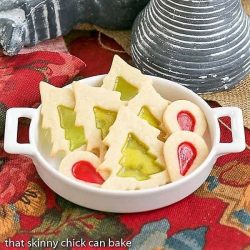 A white dish holds stained glass Christmas tree cookies with green centers and round cookies with red teardrop centers, placed on a red patterned cloth next to striped decor—one of 40 Cute Christmas Food Ideas for your holiday table.