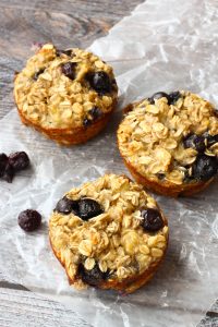 Three Baked Blueberry Oatmeal Cups with juicy blueberries rest on parchment paper, placed on a rustic wooden surface.