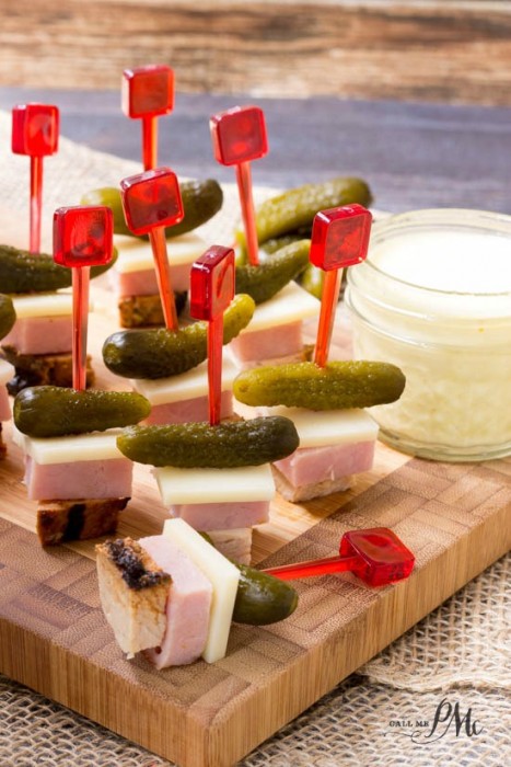A wooden board displays Cuban Sandwich on a Stick Appetizer—red toothpicks stacked with chicken, cheese, ham, and pickle slices. A jar of creamy dipping sauce sits nearby.