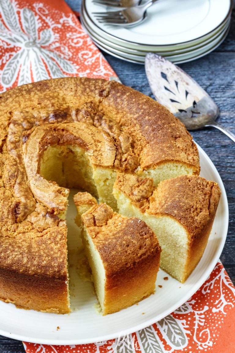 A sliced bundt Sour Cream Pound Cake Recipe on a white plate, with a serving utensil, plates, and forks in the background on a patterned cloth.