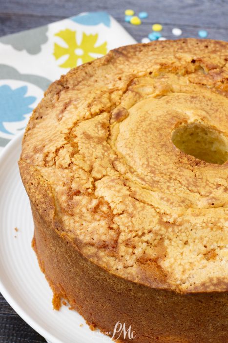 A close-up of a Million Dollar Pound Cake, golden brown with a hole in the center, sits on a white plate. A patterned napkin is partially visible in the background.