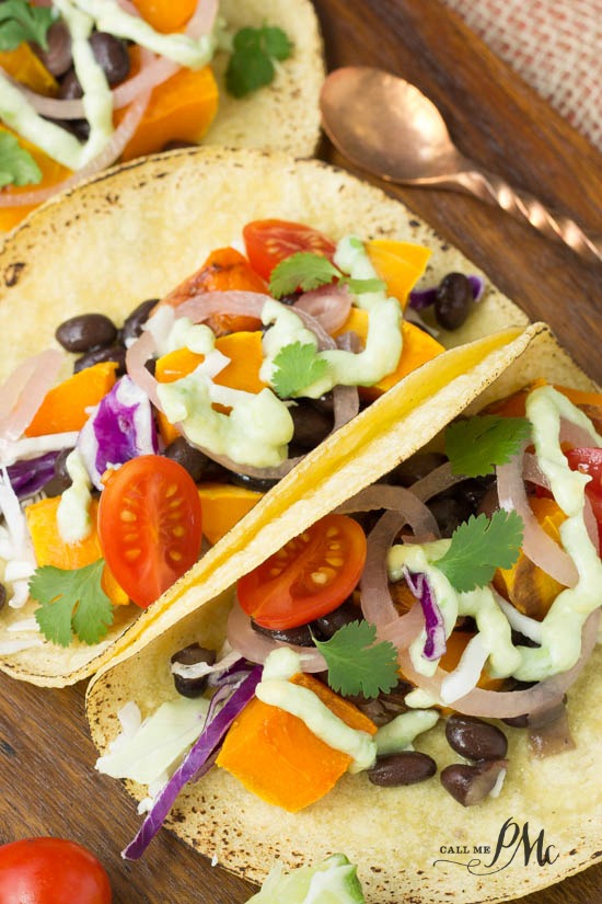Black Bean Butternut Squash Tacos with Avocado Cream Sauce recipe crisp, bright veggies are tucked in a corn tortilla for maximum flavor! Close-up of open tacos filled with cherry tomatoes, orange bell peppers, pickled onions, cabbage, cilantro, and a drizzle of green sauce on a wooden surface with a copper spoon.