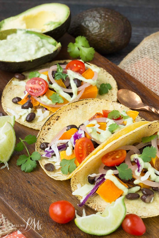 Black Bean Butternut Squash Tacos with Avocado Cream Sauce recipe tons of veggies tuckd in corn tortillas! Close-up of open tacos filled with cherry tomatoes, orange bell peppers, pickled onions, cabbage, cilantro, and a drizzle of green sauce on a wooden surface with a copper spoon.