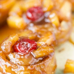 A close-up of a caramel-glazed pastry topped with a red cherry and small apple pieces evokes the indulgent allure of Pineapple Upside Down Cinnamon Rolls.