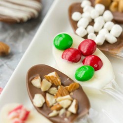 Discover the art of how to make chocolate-covered spoons, beautifully topped with almonds, red and green candies, white marshmallows, and pink sprinkles. These decorative delights are elegantly arranged on a white tray, ready to dazzle your guests.