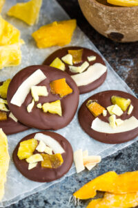 Four dark chocolate discs topped with dried pineapple and coconut flakes on parchment paper, surrounded by pieces of dried mango to create tropical dried fruit chocolate bites.