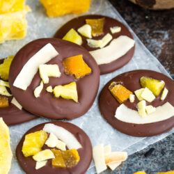 Four dark chocolate discs topped with dried pineapple and coconut flakes on parchment paper, surrounded by pieces of dried mango to create tropical dried fruit chocolate bites.