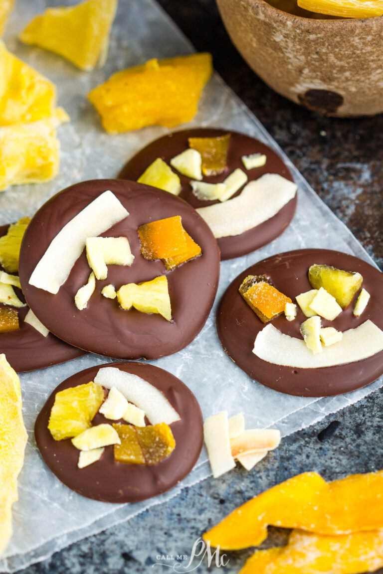 Four dark chocolate discs topped with dried pineapple and coconut flakes on parchment paper, surrounded by pieces of dried mango to create tropical dried fruit chocolate bites.