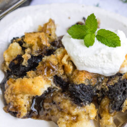 A close-up of a delectable 3 Step Oreo Cookie Dump Cobbler featuring golden cake and dark cookie pieces, topped with whipped cream and a sprig of mint, served on a white plate with a spoon.
