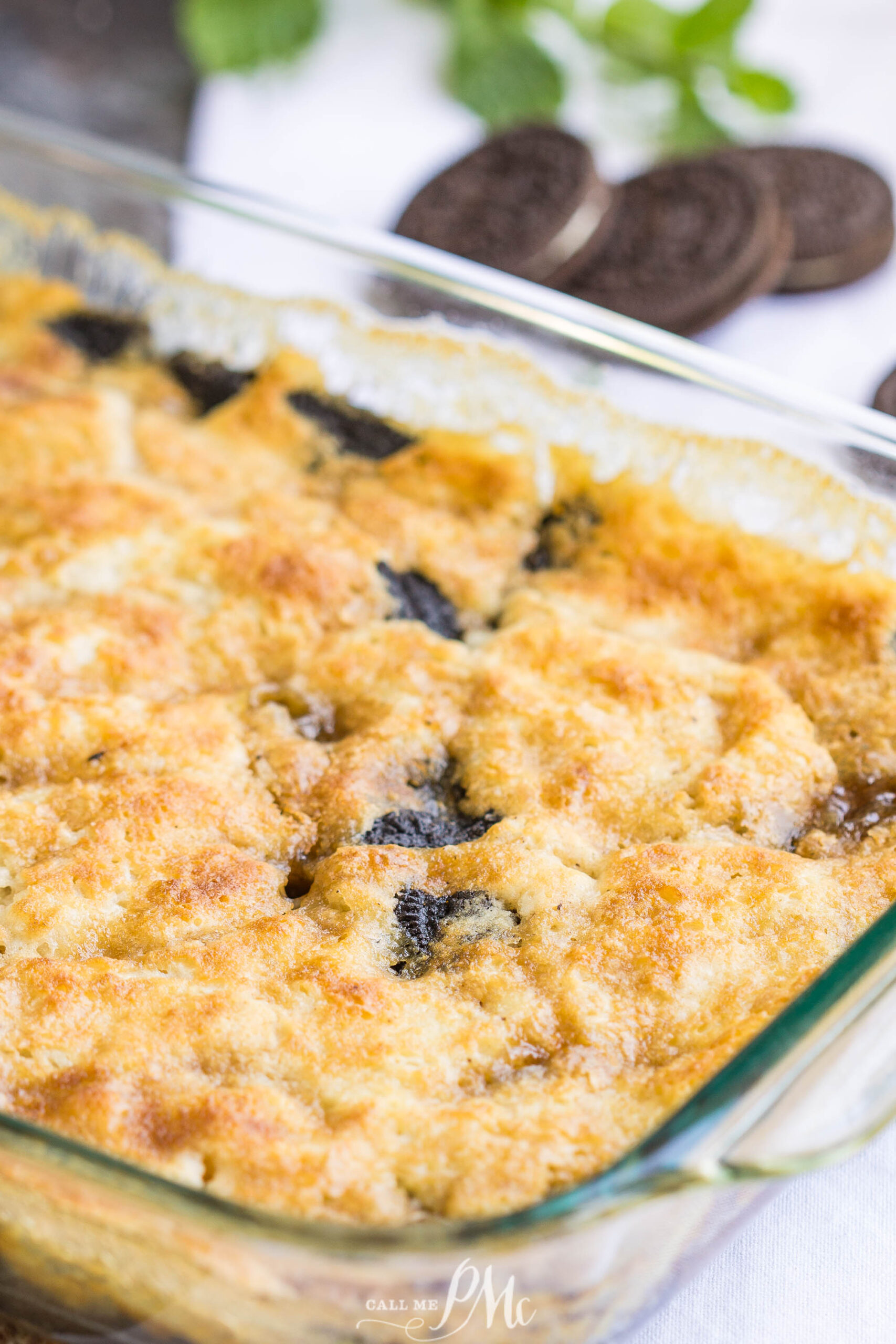 3 Step Oreo Cookie Dump Cobbler recipe. It's an easy dump-type dessert with crushed Oreo cookies A close-up of a 3 Step Oreo Cookie Dump Cobbler showcases a baked dessert with a golden, crumbly top and visible chocolate cookie pieces in a glass dish. Oreo cookies and mint leaves are blurred in the background.