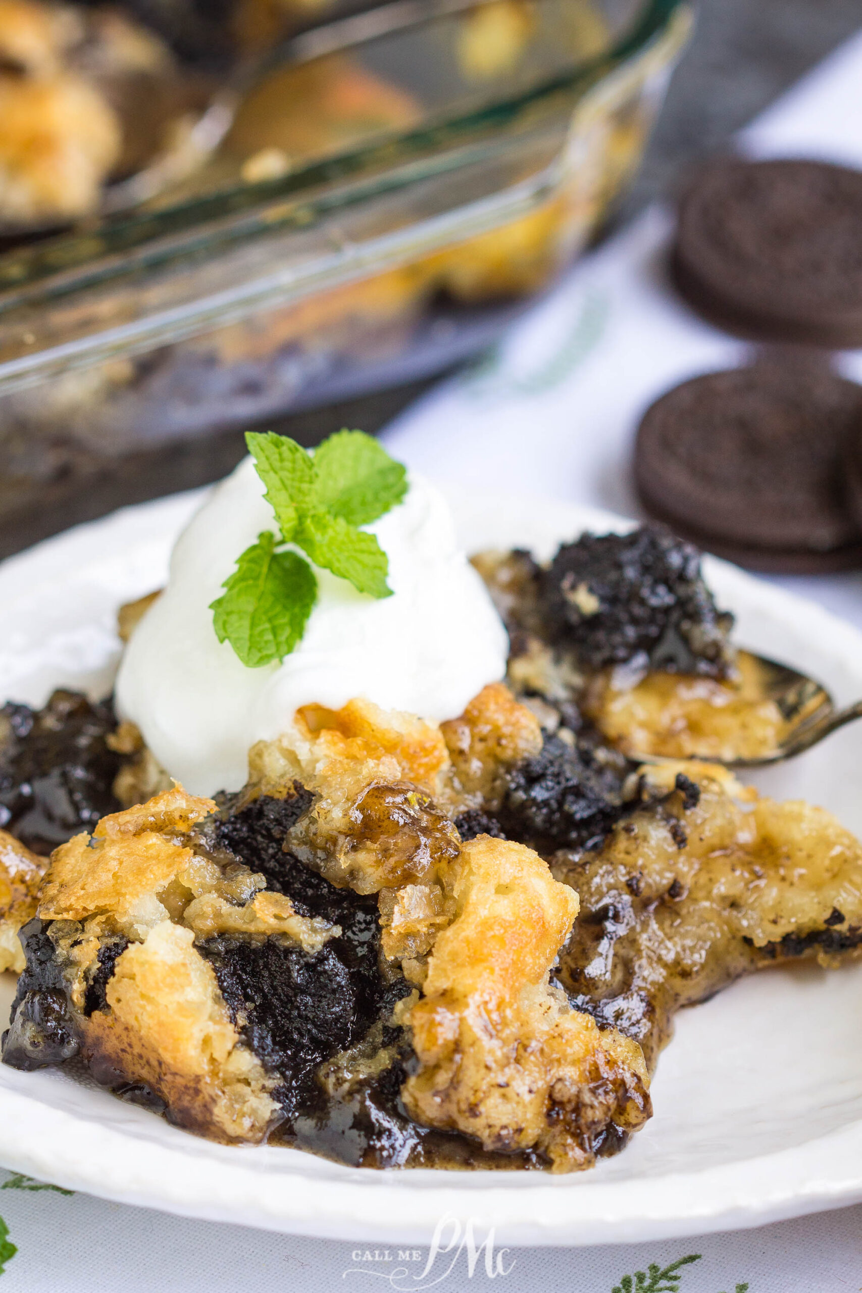 3 Step Oreo Cookie Dump Cobbler recipe. It's an easy dump-type dessert with crushed Oreo cookies A serving of 3 Step Oreo Cookie Dump Cobbler topped with whipped cream and a mint sprig is on a white plate, with more cobbler and Oreo cookies visible in the background.
