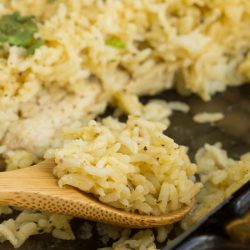 A wooden spoon scooping seasoned rice from a glass baking dish, with more rice and herbs in the background—perfect for serving this comforting No Peek Chicken Rice Recipe.