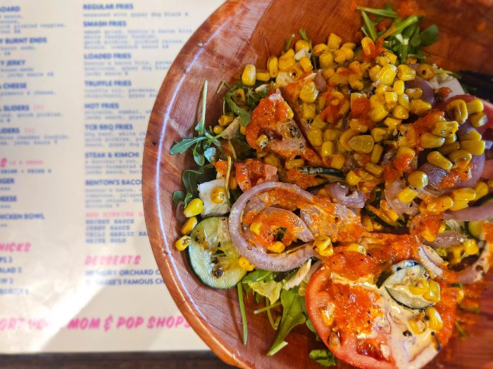 A wooden bowl of salad with grilled corn, tomato, onion, and greens sits on a table next to a blurred restaurant menu—perfect inspiration if you're searching for Where to Eat in Tupelo MS.