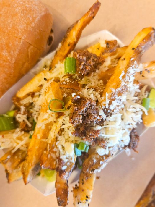 A serving of French fries topped with shredded cheese, ground meat, and chopped green onions sits next to part of a sandwich bun—a tasty option for anyone exploring Where to Eat in Tupelo MS.