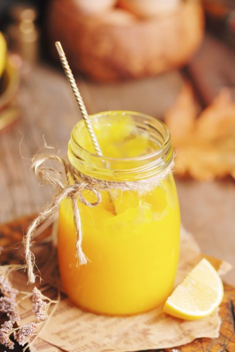 A glass jar filled with yellow homemade orange curd, tied with twine, with a metal spoon inside and a lemon wedge beside it on a rustic wooden surface.