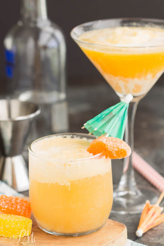 Two orange cocktails—one in a tumbler, one in a martini glass—garnished with a green paper umbrella and orange candy, with cocktail tools and candy in the background, evoke the sweetness of a Dreamsicle Martini Recipe.