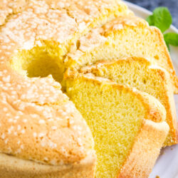 A golden, fluffy Twelve Yolk Cake with a sugar-crusted top is sliced on a white plate, revealing its soft, moist interior. Fresh mint leaves garnish the plate.