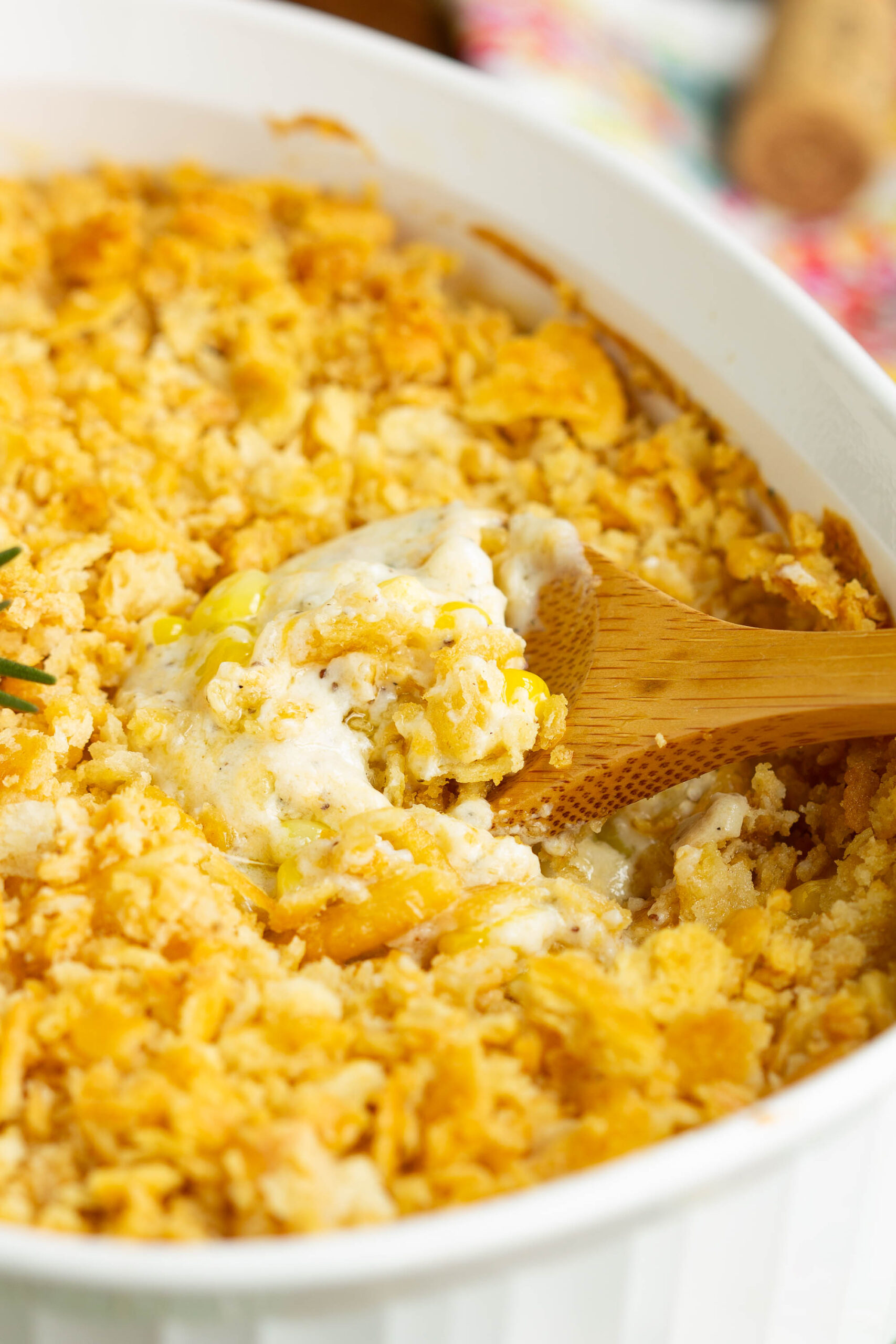 A close-up of a cheesy Baked Creamed Corn Casserole Recipe in a white baking dish with a crunchy, golden topping. A wooden spoon scoops out a portion, revealing a creamy, melted interior—made with no 'cream of' soup.
