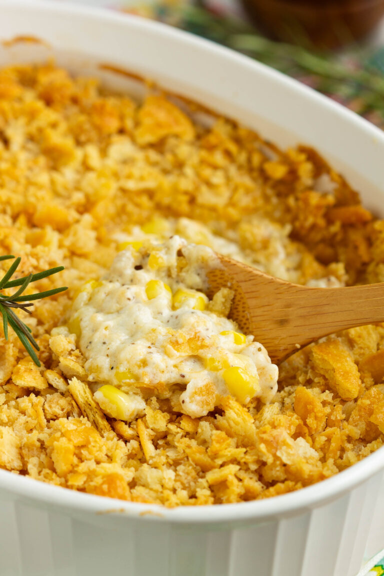 A wooden spoon scoops creamy Baked Creamed Corn Casserole Recipe with a golden, crumbly cracker topping—made with no 'cream of' soup—from a white baking dish. A sprig of rosemary rests beside the dish.