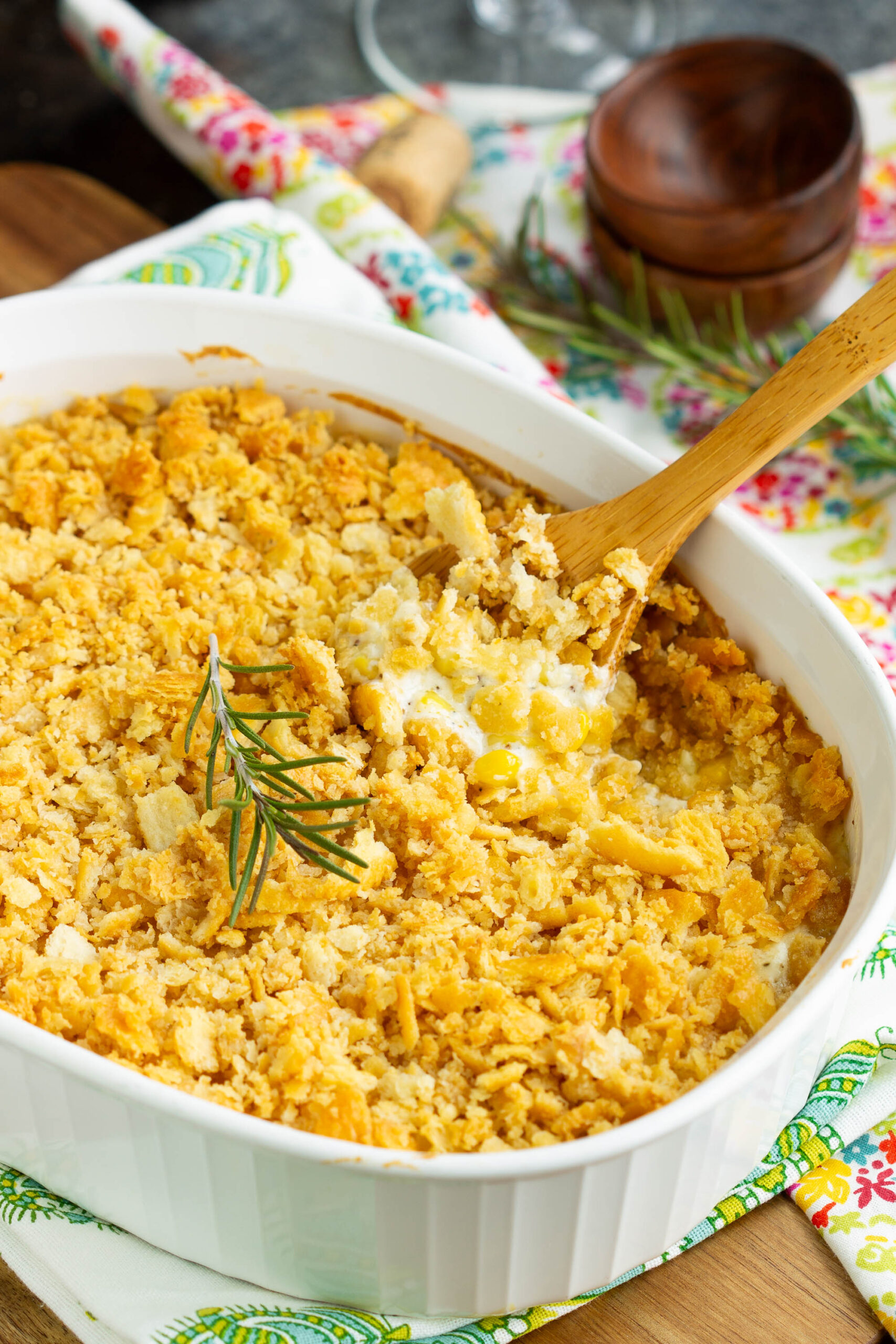 Baked Creamed Corn Casserole Recipe | No 'cream of' soup - Be a dinner-time hero when you make this delicious sweet-and-savory side dish! It's a fabulously easy recipe that is always a family favorite and a hit at pot-lucks. A white baking dish filled with Baked Creamed Corn Casserole, topped with a golden, crumbly cracker crust. A wooden spoon scoops out a serving, and a sprig of rosemary garnishes this no 'cream of' soup recipe.