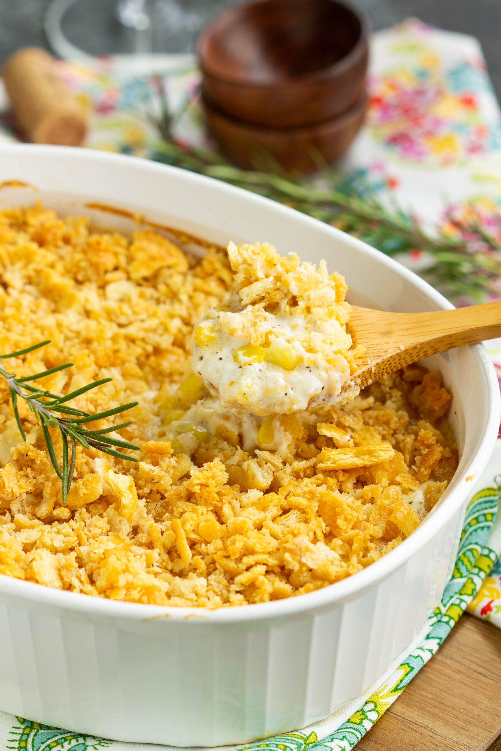 Casserole. Baked Creamed Corn Casserole Recipe | No 'cream of' soup - Be a dinner-time hero when you make this delicious sweet-and-savory side dish! It's a fabulously easy recipe that is always a family favorite and a hit at pot-lucks. A white baking dish filled with a creamy Baked Creamed Corn Casserole Recipe, topped with golden, crunchy crumbs. A wooden spoon lifts a serving, garnished with fresh rosemary. No 'cream of' soup is used. Bright, colorful napkins are in the background.