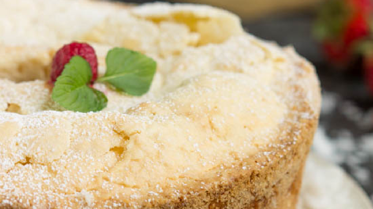 Close-up of a Coconut Cream Cheese Pound Cake, dusted with powdered sugar and topped with two mint leaves and a raspberry, with a blurred background showing part of a strawberry.