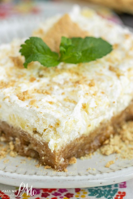 This Hawaiian Pineapple Delight Dessert Recipe is rich but light. It has a graham cracker bottom and a refreshing pineapple spiked whipped topping layer. A creamy dessert square, topped with a mint leaf and crumbled topping, sits elegantly on a white plate.