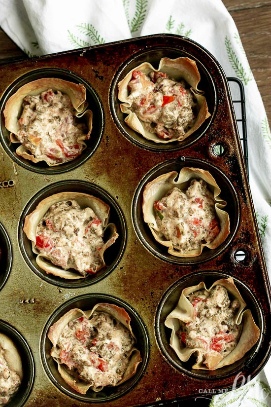 A muffin tin filled with baked phyllo cups reminiscent of Cream Cheese Sausage Rotel Wonton Cups, each containing a creamy mixture of ground meat and diced red bell peppers, placed on a wooden surface with a white cloth nearby.
