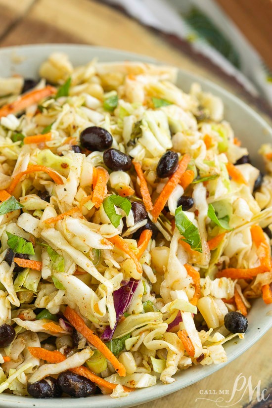 Tex Mex Coleslaw is packed with flavor and perfect for summer cookouts. It also adds a nice crunch to tacos, fajitas, and wraps. #coleslaw #salad #Mexican #TexMex A vibrant slaw sits in a bowl, featuring carrots, black beans, and shredded cabbage on a wooden board. A fork and napkin linger in the background, completing the scene.