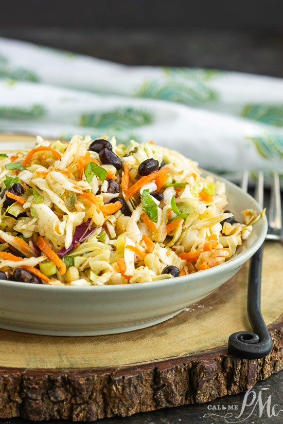 Tex Mex Coleslaw is packed with flavor and perfect for summer cookouts. It also adds a nice crunch to tacos, fajitas, and wraps. #coleslaw #salad #Mexican #TexMex A vibrant Tex Mex coleslaw sits in a bowl, featuring carrots, black beans, and shredded cabbage on a wooden board. A fork and napkin linger in the background, completing the scene.