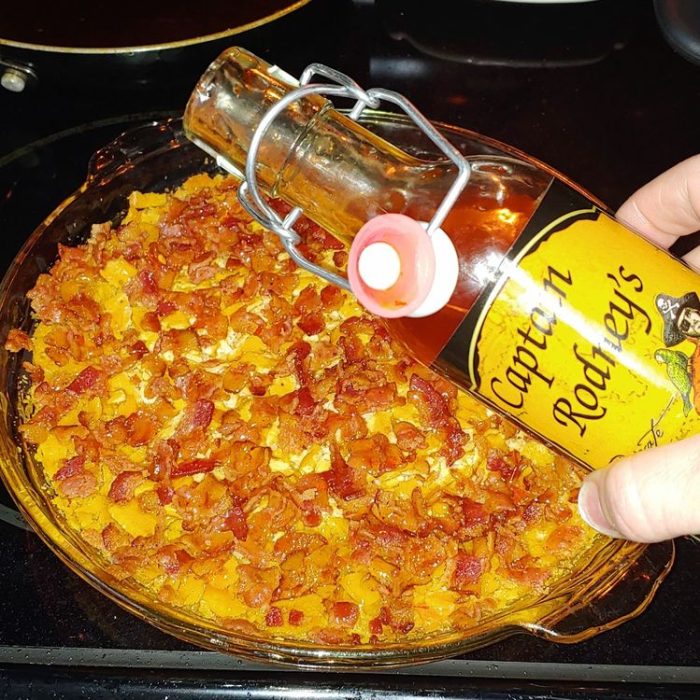A hand holds a bottle of Captain Rodney's sauce over a baked Captain Rodney's Dip, topped with chopped bacon and served in a glass pie dish.