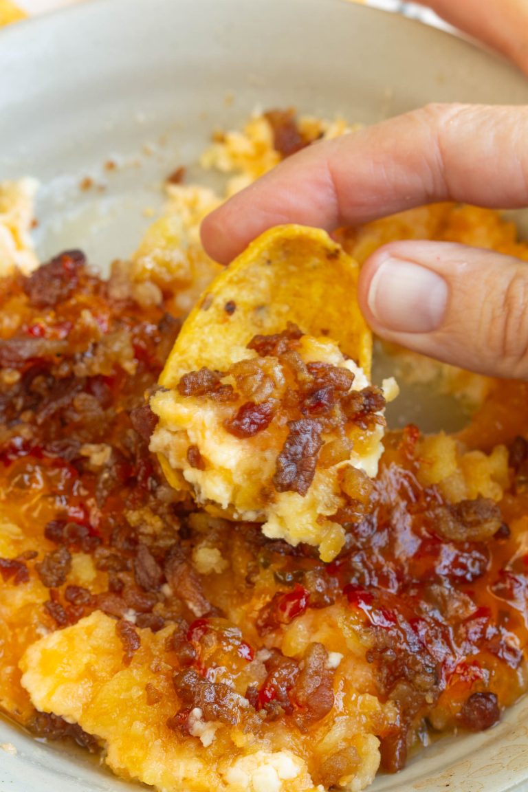 A hand holds a chip scooping Captain Rodney's Dip, a cheesy dip topped with bacon bits and a red sauce, from a bowl.