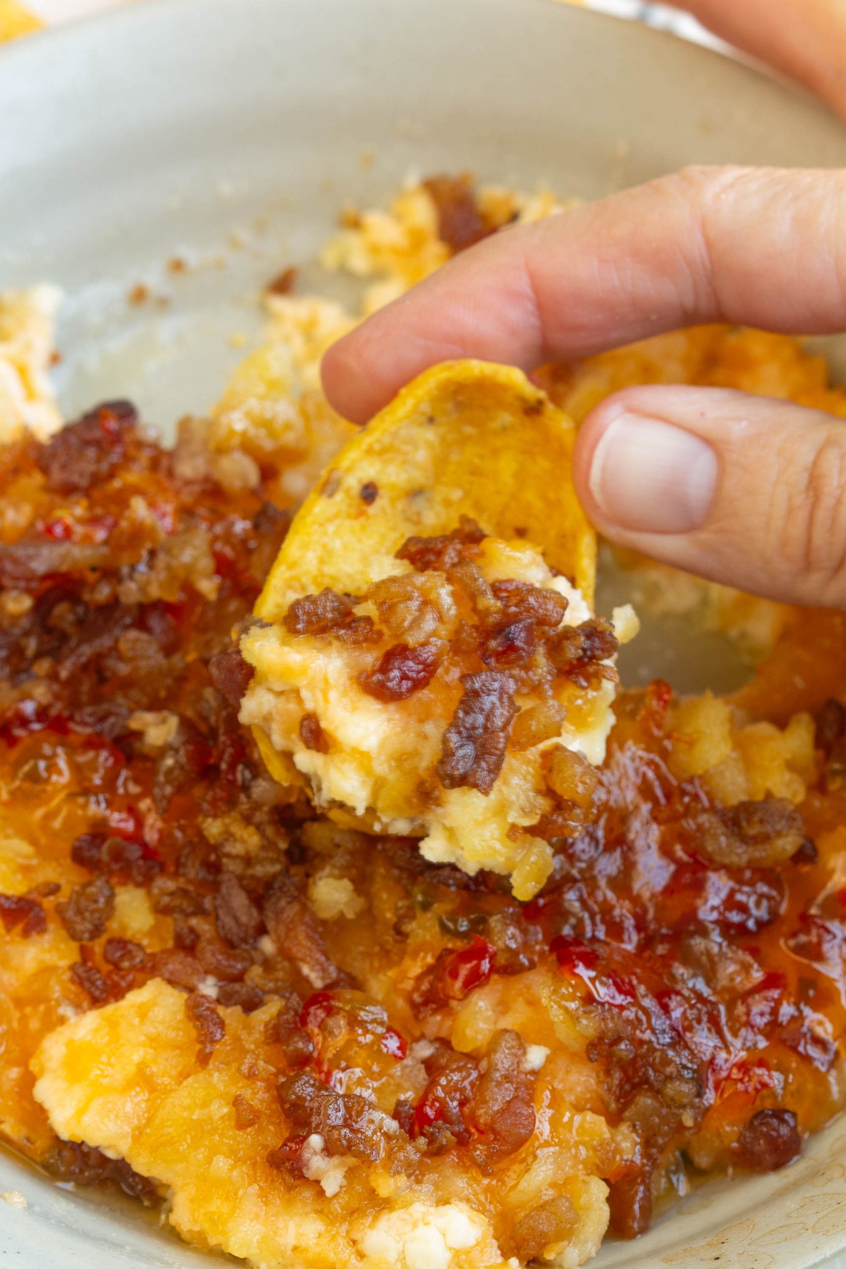 Captain Rodney's Dip is the ultimate party food. This outrageously good dip recipe is what every party, tailgate, and cookout needs! #dip #appetizer #creamcheese #CaptainRodneys #bacon #tailgating #watchparty #easy A hand holds a chip scooping Captain Rodney's Dip, a cheesy dip topped with bacon bits and a red sauce, from a bowl.