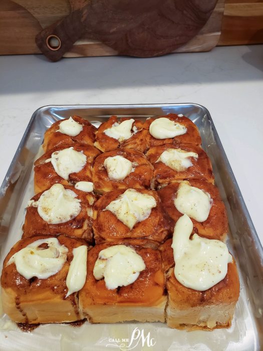 A metal tray filled with twelve Quick Cinnamon Hawaiian Rolls topped with dollops of cream cheese frosting, set on a white countertop.