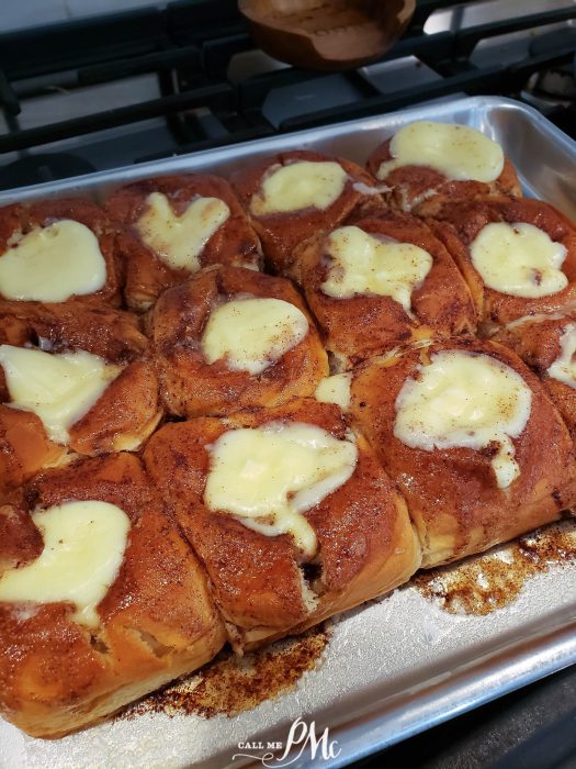 A baking tray of rolls topped with dollops of cream cheese frosting, fresh out of the oven and set on a stovetop.