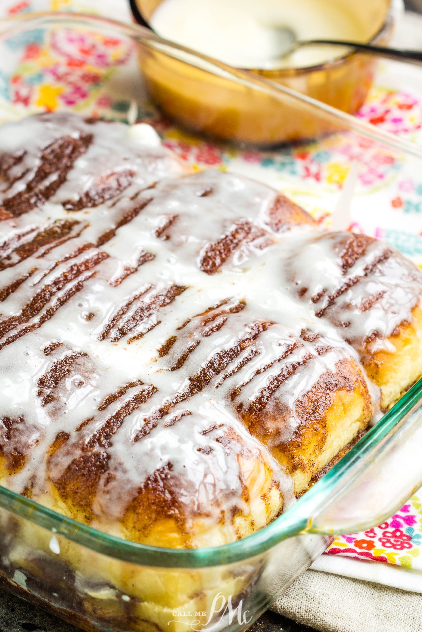 A glass baking dish filled with Quick Cinnamon Hawaiian Rolls topped with white icing, resting on a colorful patterned cloth.