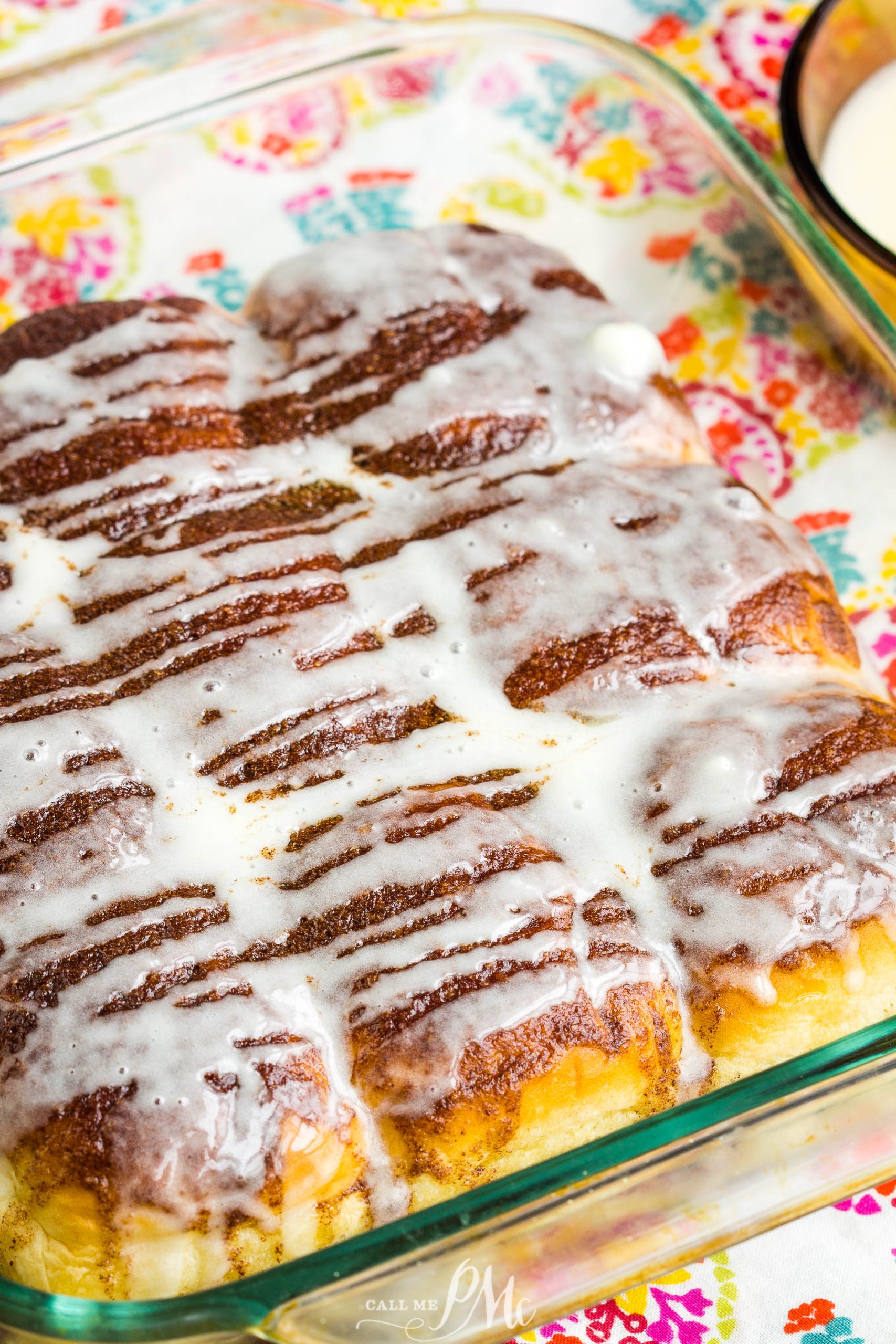 A glass baking dish filled with Quick Cinnamon Hawaiian Rolls topped with white icing, placed on a colorful patterned surface.