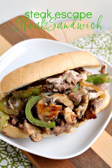 A Grand Escape Steak Sandwich on a hoagie roll filled with sliced steak, green bell peppers, mushrooms, and melted cheese is served on a white plate. The words "steak escape Steak Sandwich" are written above the sandwich.