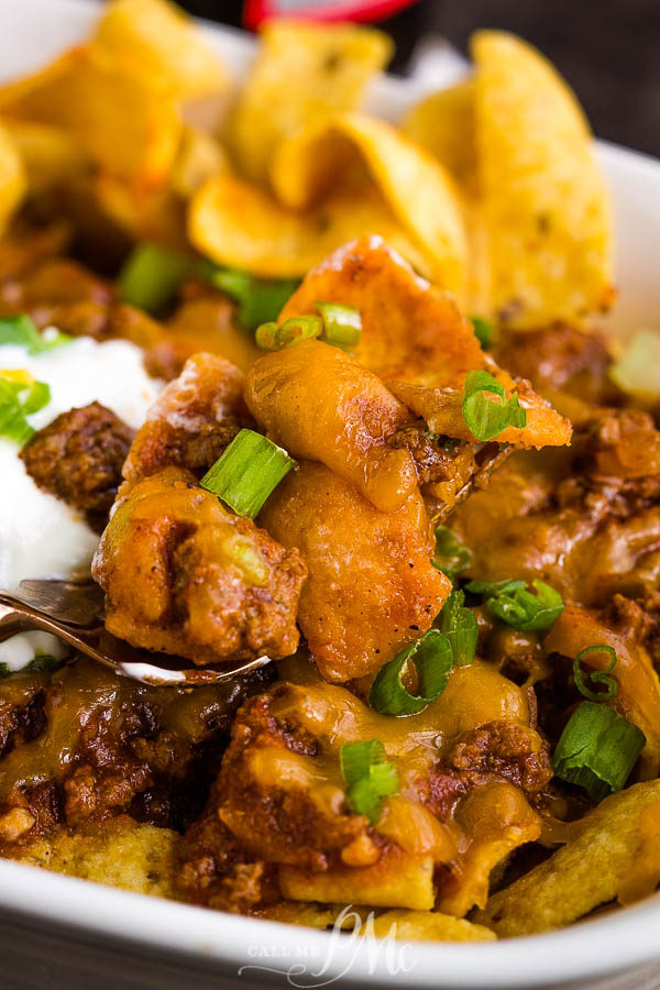 Frito Chili Pie Recipe is classic comfort and gameday food. It's simple, hearty, delicious, takes just minutes to make, and the whole family loves it! #cornchips #Fritos #chips #chili #cheese #cheddar #meal #fastfood #copycat Close-up of a Frito Chili Pie featuring seasoned meat, melted cheese, diced green onions, and a dollop of sour cream on top of corn chips.